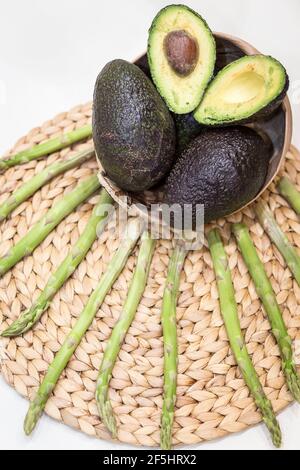 Vertikale Draufsicht auf geschnittene und vollreife Avocados, die auf einem Korb unter Palmfaser matt angeordnet sind und mit rohen grünen Spargeln dekoriert sind Stockfoto