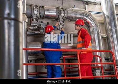 Young Worker in Red Coveralls und HardHat stehen neben Maintenance Worker in blauer Uniform. Werksinneneinrichtung mit großen Rohrleitungen. Stockfoto