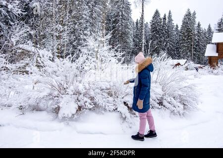 Eine junge Frau in warmer Winterkleidung spaziert zwischen den schneebedeckten Büschen und Bäumen auf dem Land. Stockfoto