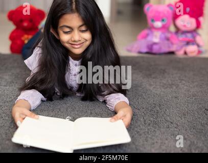 Porträt eines niedlichen indischen Schulmädchen entspannen auf einer Spielmatte, lächeln und ein Tagebuch Buch von Spielzeug umgeben lesen. Stockfoto