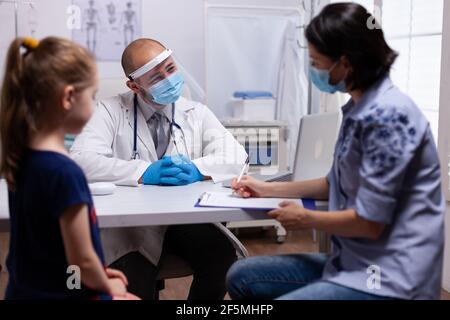 Mutter Ausfüllen des medizinischen Fragebogens in der Arztpraxis in der Klinik. Kinderarzt mit Schutzmaske und Visier gegen covid19 sprechen über diagnostische Ergebnisse Bereitstellung von Versorgungsleistungen Stockfoto