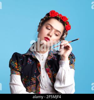 Junge Frau im Bild der mexikanischen Künstlerin Frida mit roten Rosen im Haar. Mit einem Pinsel in den Händen, berühren Augenbrauen. Stockfoto