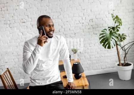 Glücklicher Geschäftsmann, der im Heimbüro steht, ein Telefongespräch mit einem Kollegen führt und ein Projekt diskutiert. Stilvoller afroamerikanischer Mann im Gespräch Stockfoto