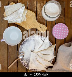 Wonton in Dreiecksform vorbereiten. Wonton auf einem Teller auf einem dunklen Holztisch, mit etwas Mehl und Wasser in kleinen Schüsseln sowie Knödelverpackungen. Stockfoto