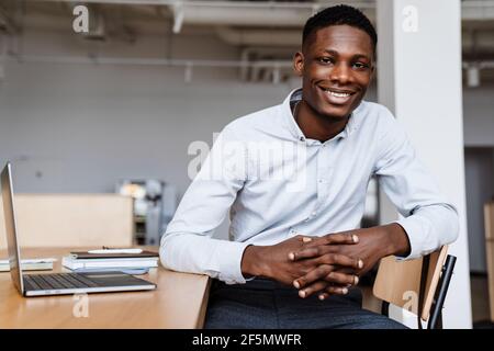 afroamerikanischer glücklicher Mann, der die Kamera anlächelt und mit ihm arbeitet Laptop im Büro Stockfoto