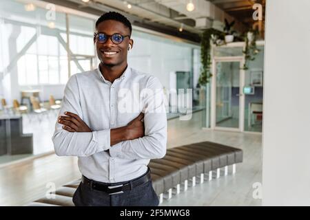 afroamerikanischer junger Mann in Brillen und Ohrhörern, die drinnen stehen Während der Arbeit im Büro Stockfoto