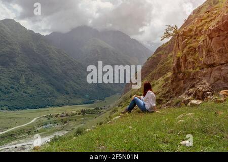 Eine Frau mit roten Haaren in einer weißen Bluse und Jeans sitzt vor einer Berglandschaft, getönt Stockfoto