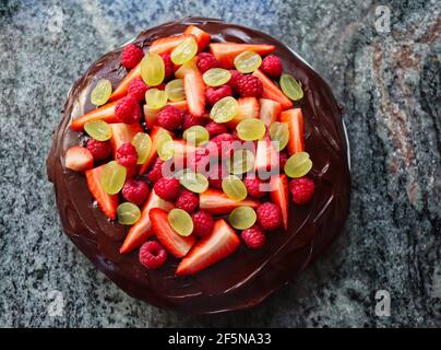 Dunkler Schokoladenkuchen mit Obst von oben auf der Küchenunterseite aus Marmor. Tarte mit Schokoladenglasur und Erdbeeren, Trauben und Himbeeren. Stockfoto