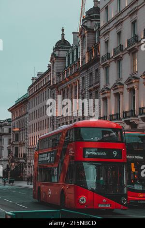 London UK Februar 2021 Vertikale Aufnahme von zwei leuchtend roten Doppeldeckerbussen, die spät am Tag durch London fahren. Die Gebäude im Hotel strahlen ein unglaubliches Licht aus Stockfoto