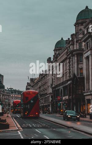 London UK Februar 2021 Vertikale Aufnahme von zwei leuchtend roten Doppeldeckerbussen, die spät am Tag durch London fahren. Die Gebäude im Hotel strahlen ein unglaubliches Licht aus Stockfoto