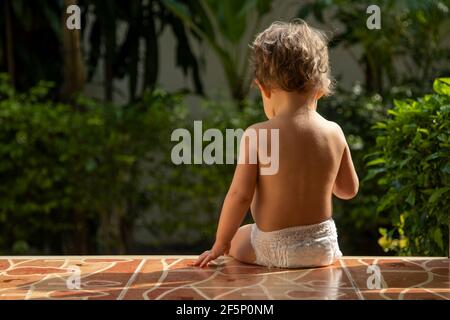 Ein charmantes Kleinkind sitzt auf den Stufen eines Hauses im Sonnenlicht. Rückansicht Stockfoto