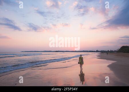 Eine junge Touristenin, die auf einen Spaziergang entlang der Strand bei einem bunten pastellfarbenen Sonnenuntergang am Urlaubsziel Weligama Beach an der Südküste Stockfoto