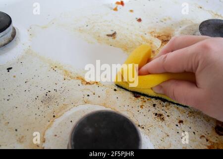 Eine Hand mit einem gelben Waschschwamm wäscht die sehr schmutzige fettige Oberfläche des Gasherd. Nach dem Schwamm bleibt eine saubere Spur. Stockfoto