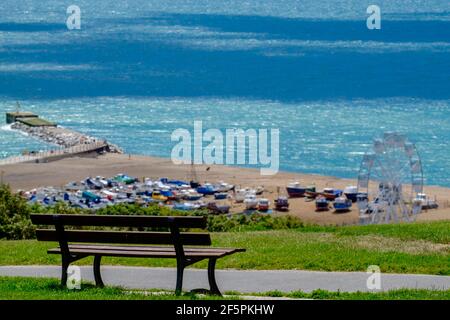 Staycation Idee. Eine leere Bank mit Blick auf den Ärmelkanal und den verschwommenen Stade Beach mit dem Big Wheel und befahrene Fischerboote. Hastings. Stockfoto