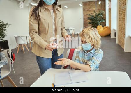 Ausgeschnittene Aufnahme einer jungen Lehrerin mit einem Alkoholspray Zum Desinfizieren der Hände der Schüler im Klassenzimmer Stockfoto