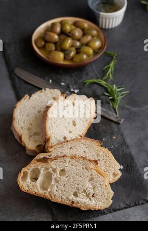 Italienisches Ciabatta-Brot auf Schneidebrett mit Kräutern, nativem Olivenöl extra und marinierten Oliven auf dunklem Grunge-Hintergrund Stockfoto