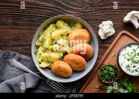 Gebratener Blumenkohl- und Käsebroketten mit gekochten Kartoffeln und sauer Creme Dip auf rustikalem Holzhintergrund Stockfoto