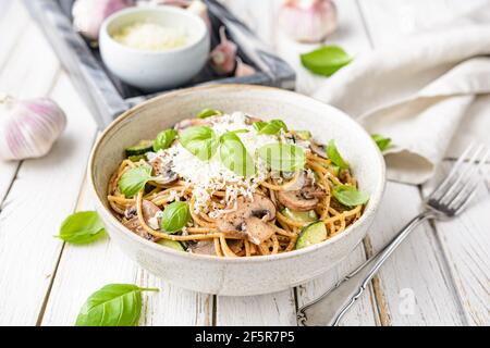 Gesunde Vollkornspaghetti mit sautierten Pilzen Knoblauch und Zucchini, gekrönt mit geriebenem Käse und Basilikumblättern auf rustikalem Holzhintergrund Stockfoto