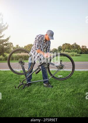 In voller Länge Schuss von jungen Mann in Casual tragen Befestigung Pedale auf seinem Fahrrad, auf dem Rasen im öffentlichen Park stehen Stockfoto