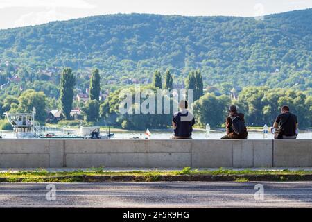 Rückansicht von drei Touristen auf Beton-Hochwasserschutzzaun neben der Donau sitzen, Wald auf der anderen Seite, Visegrad, Ungarn Stockfoto