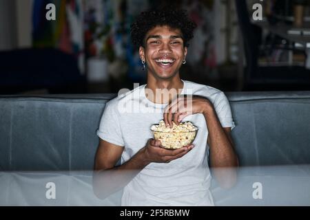 Fröhlicher junger Mann im Schlafanzug, der Popcorn aus einer Schüssel nimmt, während er sich Komödie ansieht und zu Hause auf einer bequemen Couch sitzt Stockfoto