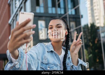 Schöne afroamerikanische Frau mit stilvollen Frisur unter Selfie, zeigt Siegeszeichen. Influencer, der Video-Streaming über Mobiltelefone nutzt Stockfoto