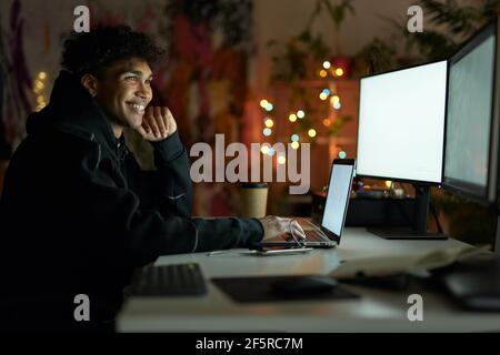 Seitenansicht des glücklichen jungen Kerl mit Piercing sitzen an Der Tisch vor vielen Computer-Monitore und arbeiten Von zu Hause spät am Abend Stockfoto