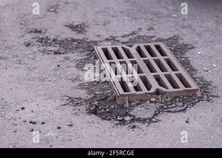 Regenwasserrost im Asphalt. Ein altes Regengitter ragt aus dem Asphalt. Stockfoto