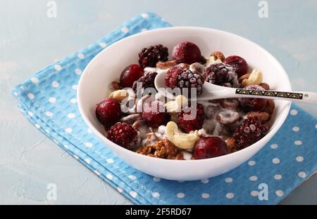 Müsli knusprig mit Naturjoghurt, gefrorenen Kirschen und Brombeeren, Schokolade und Nüssen in weißer Schale auf blauem Hintergrund. Stockfoto