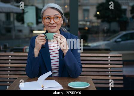 Mist Geschäftsfrau trinkt Kaffee, sitzt im Café. Porträt der nachdenklichen asiatischen Frau trägt stilvolle Brillen, hält Tasse Tee Stockfoto