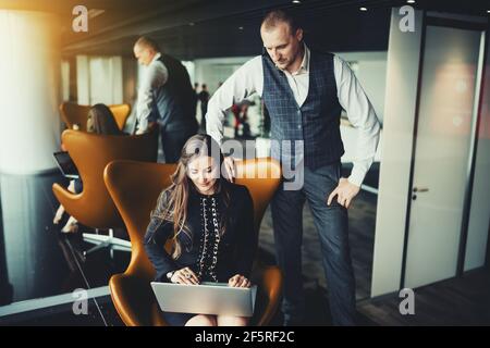 Eine herrisch reife Geschäftsfrau mit langen Haaren sitzt darauf Ein orangefarbener Sessel und mit ihrem Laptop, während ihr Mann Die Kollegin steht neben ihr und Stockfoto