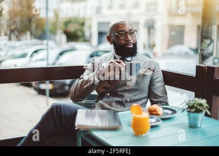 Ein ausgefallener bärtiger, haarloser Senior Unternehmer in Gläsern und einem stilvollen Maßkostüm sitzt morgens in einem Outdoor-Café und trinkt einen Stockfoto