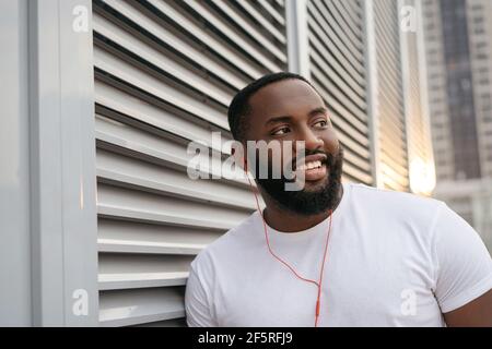 Authentisches Porträt eines schönen afroamerikanischen Mannes, der für Bilder posiert, lächelt und auf der Straße steht. Fröhlicher bärtiger Kerl, der draußen Musik hört Stockfoto