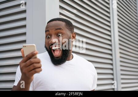 Aufgeregt afroamerikanischen Mann mit Handy, Online-Shopping. Portrait von überglücklich online Lotterie Gewinner Blick auf digitalen Bildschirm Stockfoto