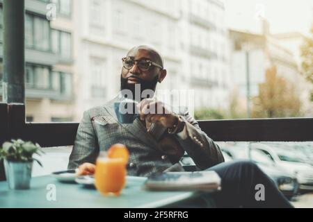 Das Porträt eines gutaussehenden stilvollen wohlhabenden afrikanischen Kerl mit einem schönen schwarzen Bart, in einer Brille, kahl, in einem eleganten Anzug, sitzt an einem regnerischen Morgen Stockfoto