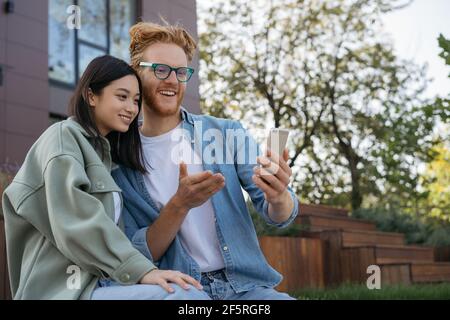 Zwei lächelnde Freunde, die online über das Mobiltelefon einkaufen. Schöne asiatische Frau und hübscher kaukasischer Mann kommunizieren miteinander und sehen sich Videos an Stockfoto