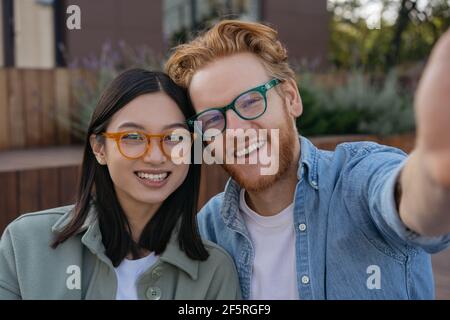 Junge lächelnde Freunde tragen stilvolle Brillen, die Selfie auf der Straße machen. Schöne asiatische Frau und hübscher kaukasischer Mann, der die Kamera anschaut Stockfoto