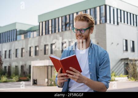 Schöner lächelnder Mann, der im Freien Buch liest. Erfolgreiche Studenten tragen stilvolle Brillen studieren, Prüfungsvorbereitung, Bildungskonzept Stockfoto