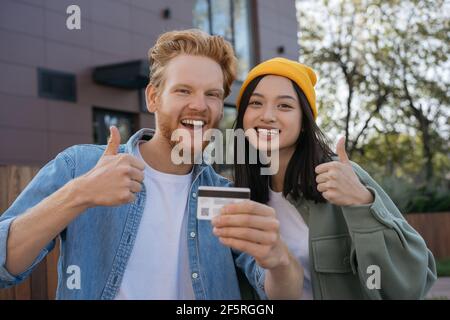 Ein paar lächelnde Freunde gemischter Rennen, die die Kreditkarte vorführen Daumen nach oben und Blick auf die Kamera Stockfoto