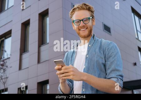 Hübscher lächelnder rothaariger Mann, der die mobile App zum Online-Shopping nutzt, um die Kamera zu betrachten. Porträt eines jungen erfolgreichen Freiberuflers mit Brille Stockfoto