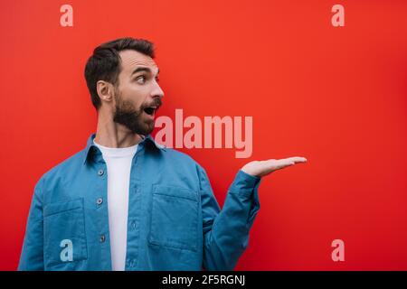 Emotionaler bärtiger Mann mit lässigem Hemd, der mit der Hand zeigt, isoliert auf rotem Hintergrund. Platz kopieren, Werbekonzept Stockfoto