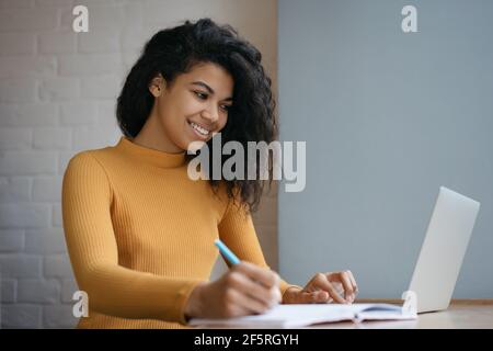 Afroamerikanische Studentin, die studiert, mit einem Laptop-Computer unterwegs ist, Notizen auf einem Notebook macht. Online-Bildungskonzept. Junge schöne Frau, die von zu Hause aus arbeitet Stockfoto