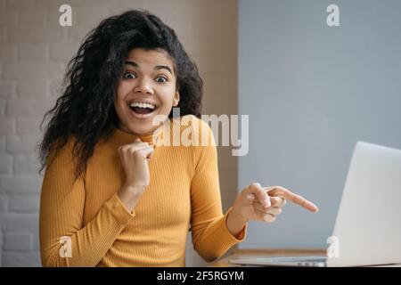 Überrascht afroamerikanische Frau zeigt Finger auf Laptop-Bildschirm. Porträt eines begeisterten erfolgreichen Influencers, der mit offenem Mund die Kamera anschaut Stockfoto