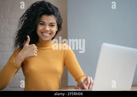 Afroamerikanische Frau, die einen Laptop benutzt, den Daumen nach oben zeigt, die Kamera anschaut und lächelt und von zu Hause aus arbeitet. Glücklicher Student beim Studieren Online Stockfoto