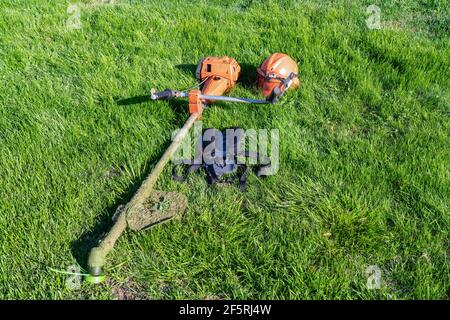 Vorderansicht des tragbaren Benzinrasenmähers, Schutzhelm mit Augen- und Gehörschutz, gepolsterte Schultergurte links nach der Arbeit auf grünem Gras la Stockfoto