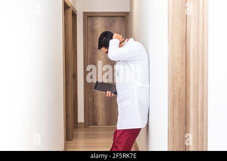 Lateinischer Arzt mit einem weißen Mantel und einem Stethoskop, das eine Tablette müde hält und sich an eine Wand im Flur eines Krankenhauses lehnt, Konzept der Gesundheitsarbeit Stockfoto