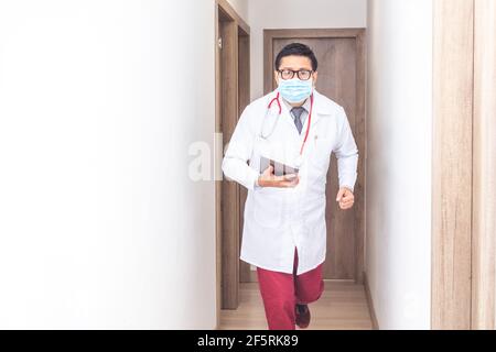 Porträt eines hispanischen Arztes mit weißem Mantel, Krawatte, Maske und Stethoskop, der in einem Krankenhausflur läuft und eine Tablette in den Händen hält. Konzept o Stockfoto