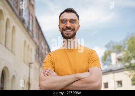 Porträt eines gutaussehenden lächelnden Mannes, der auf die Kamera schaut und auf der Straße steht. Junger selbstbewusster Geschäftsmann mit gekreuzten Armen und stylischen Brillen Stockfoto