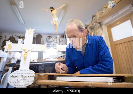 26. März 2021, Sachsen, Neudörfel: Nikolaus Dürlich schreibt in seiner Werkstatt mit einem Pinsel einen Namen auf eine Holzplatte. Der sorbische Holzbildhauer ist die dritte Generation, die Kreuze und religiöse Figuren herstellt und restauriert. Foto: Sebastian Kahnert/dpa-Zentralbild/dpa Stockfoto