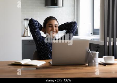 Lächelnde indische Frau, die auf den Laptop-Bildschirm schaut und sich die Hände streckt Stockfoto